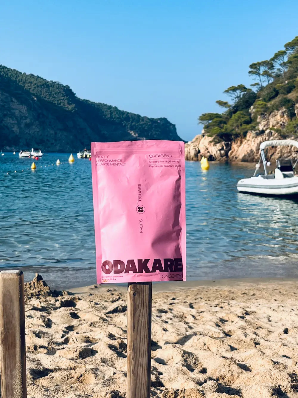 Pink Creagen+ Red Berries pouch in its original design, placed on a wooden post in front of a sandy cove with turquoise water and boats at anchor