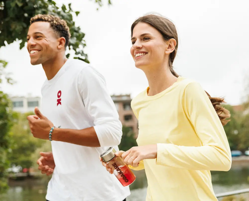 A smiling man and woman jogging by the water in the city, the woman holding an Odakare shaker filled with a red drink