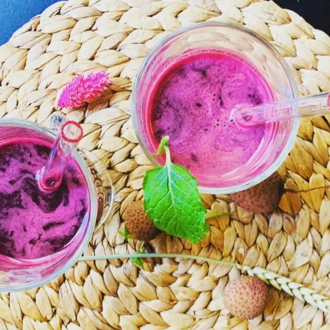 Deux verres de smoothie rose-violet avec pailles en verre, vus du dessus sur un set de table tressé, entourés de litchis, d’une feuille de menthe et d’une fleur rose