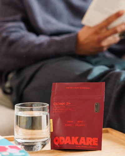 Sachet ODAK 3+ d’Odakare et verre d’eau posés sur un plateau en bois, devant une personne lisant dans son canapé, en arrière-plan flou