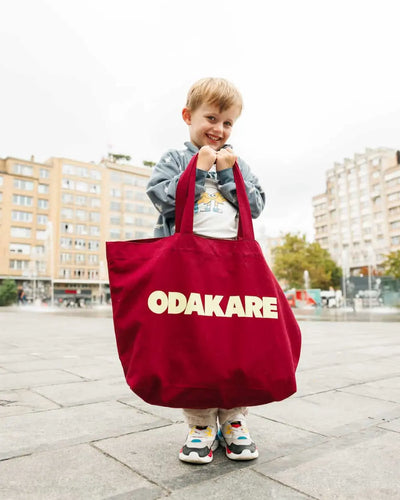 Petit garçon souriant tenant un grand tote bag bordeaux Odakare presque aussi grand que lui, sur une place en ville
